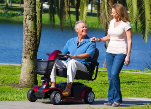 Man using a mobility scooter with a woman assisting him in a park setting.